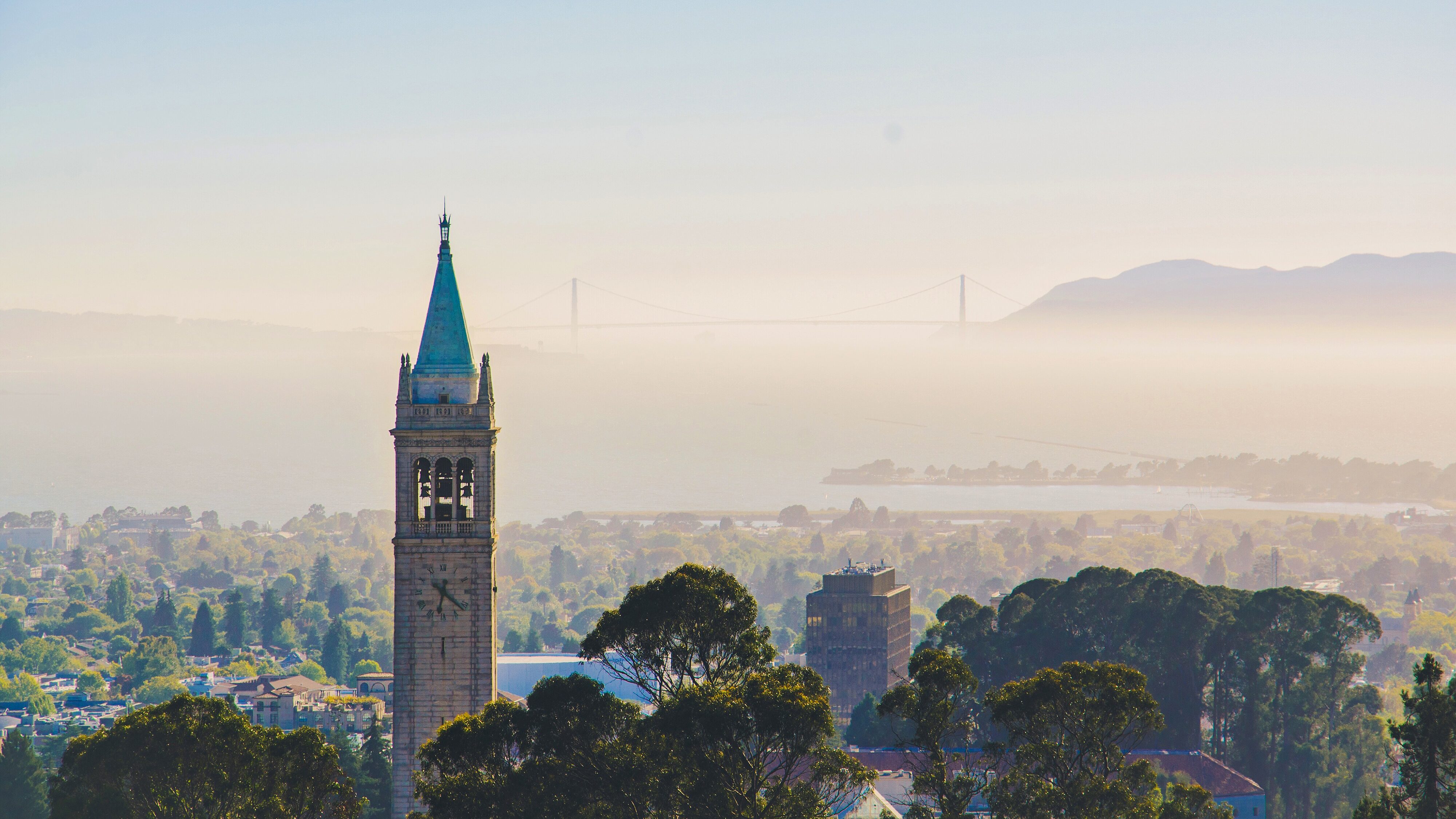 Campanile in foreground with hazy morning fog in background across the bay and partly covering Golden Gate bridge. Sunny, clear sky. UC Berkeley logo placed in top left corner.