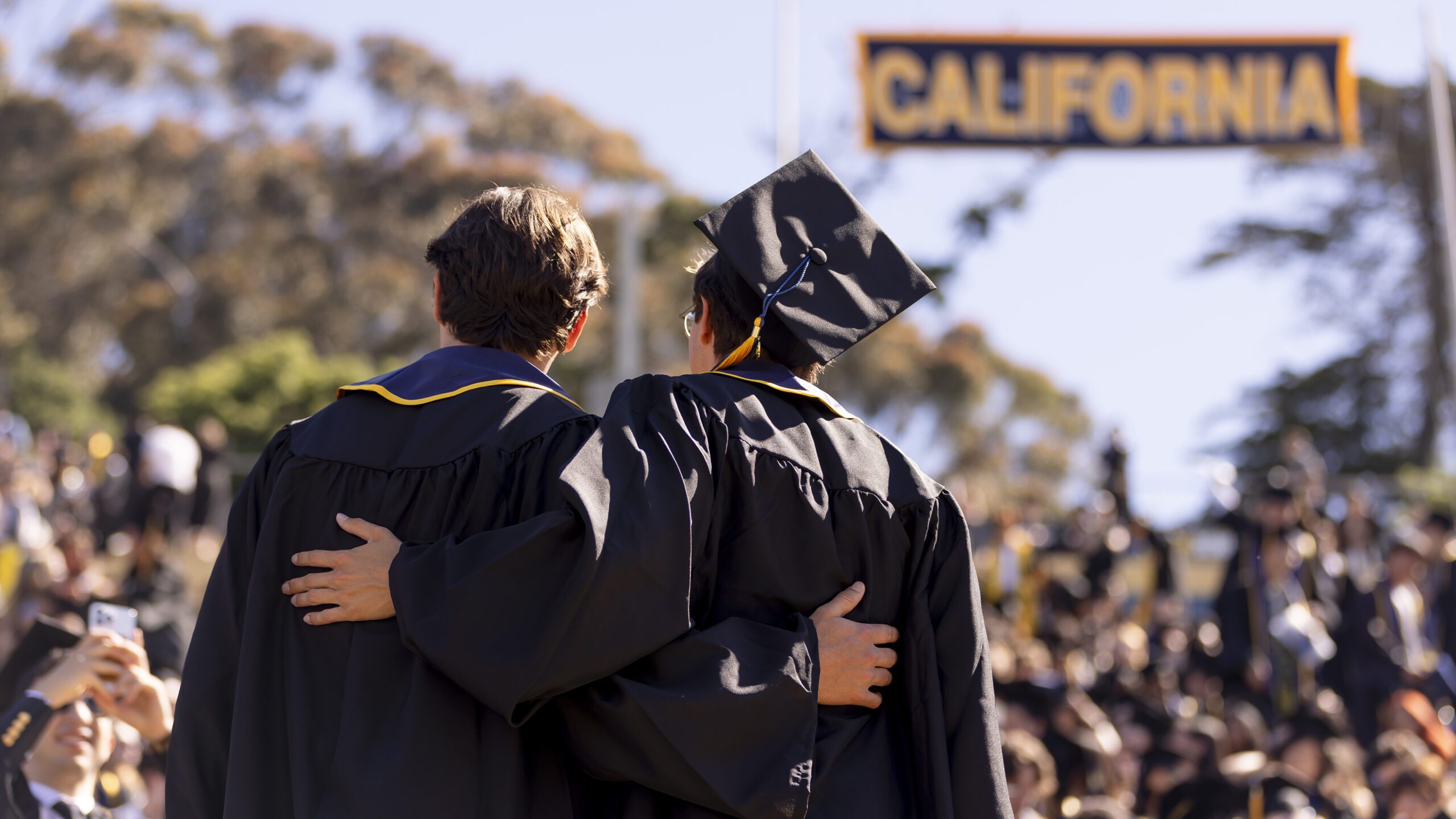 Two graduates dressed in regalia wrap their arms around each other and look out at the crowd at Commencement