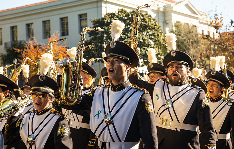 A group of 10-20 Band members in Cal uniforms hold up their Saxophones. Blurry trees and building in background. Daytime, sunny.