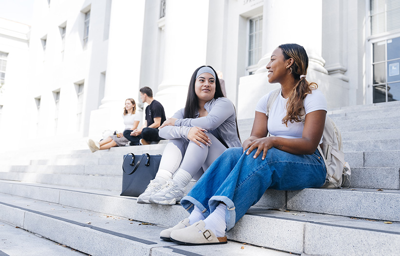 4 people sit on steps during daytime with out of focus white building behind them.